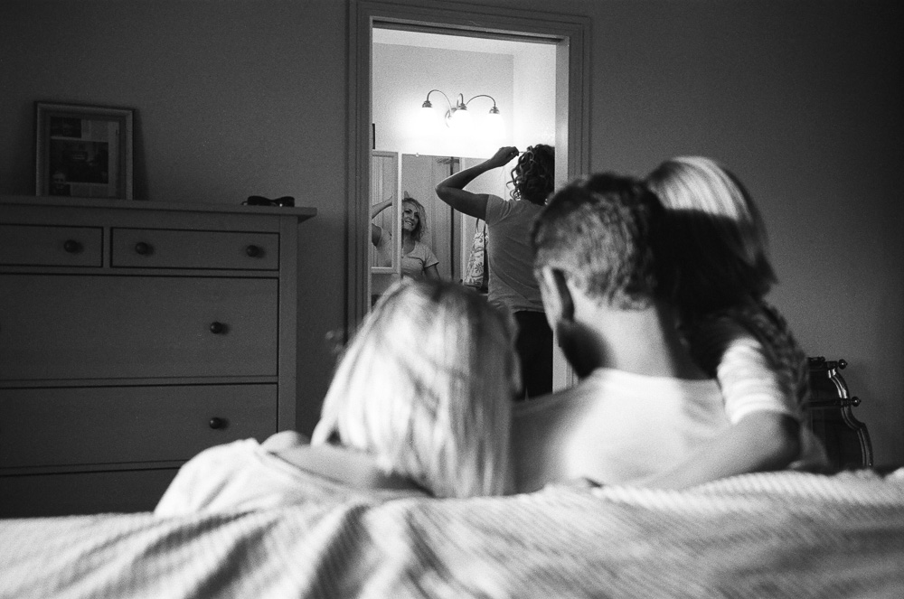 Woman (Morgan) perfecting her hair in bathroom as dad (Jeff) and daughters (Kate and Alex) wait. ©William Bay Photographic Arts.