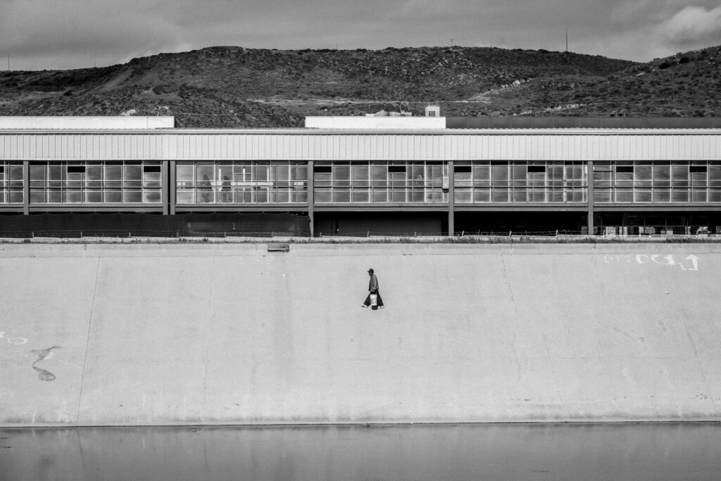 A homeless man walks along the bank to his outlet with a 5 gallon bucket.